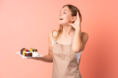 Ukraynalı aşçı pembe arka planda izole edilmiş bir kek tutuyor ve bir şeyler dinliyor.