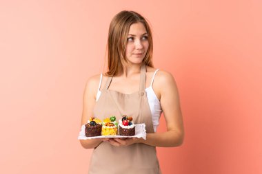 Ukraynalı pasta şefi pembe arka planda izole edilmiş bir kek tutuyor ve yan tarafa bakıyor.