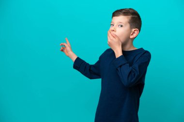 Küçük çocuk mavi arka planda izole edilmiş, sürpriz bir ifadeyle işaret ederken