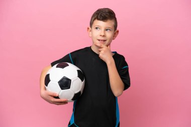 Küçük beyaz futbolcu çocuk pembe arka planda izole edilmiş, yan tarafa bakıyor ve gülümsüyor.