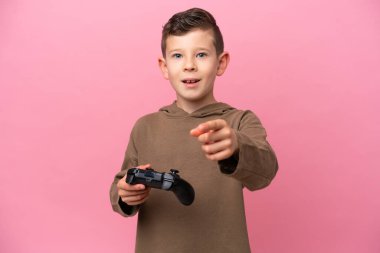 Pembe arka planda izole edilmiş bir bilgisayar oyunu kontrolörüyle oynayan küçük beyaz çocuk şaşırmış ve ön tarafı işaret ediyor.
