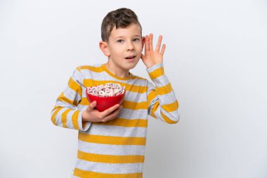 Beyaz arka planda izole edilmiş mısır gevreği kasesi tutan küçük beyaz bir çocuk kulağına elini sürerek bir şeyler dinliyor.