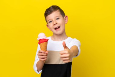 Kornet dondurması olan küçük beyaz bir çocuk sarı arka planda izole edilmiş, başparmakları havada çünkü iyi bir şey oldu.