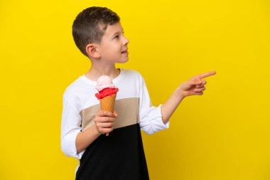 Kornet dondurması olan beyaz bir çocuk sarı arka planda izole edilmiş bir şekilde parmak ucunda bir ürün sunuyor.