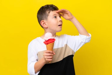 Sarı arka planda izole edilmiş kornet dondurması olan küçük beyaz çocuk yan tarafa bakarken sürpriz jest yapıyor.