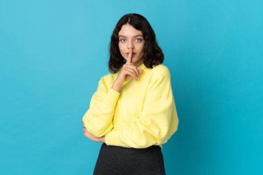 Mavi arka planda izole edilmiş genç Ukraynalı kız parmağını ağzına koyan sessizlik işareti gösteriyor.