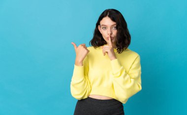 Genç Ukraynalı kız, mavi arka planda izole edilmiş, kenarı işaret ediyor ve sessizlik hareketi yapıyor.