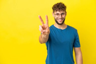 Sarı arka planda izole edilmiş yakışıklı beyaz adam gülümsüyor ve zafer işareti gösteriyor.