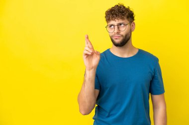 Sarı arka planda izole edilmiş yakışıklı beyaz bir adam parmaklarını çapraz yapıp en iyisini diliyor.