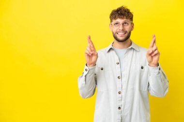 Sarı arka planda izole edilmiş yakışıklı beyaz bir adam parmaklarını çapraz yapıp en iyisini diliyor.