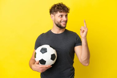 Sarı arka planda izole edilmiş yakışıklı genç futbolcu harika bir fikre işaret ediyor.