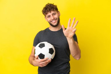 Yakışıklı genç futbolcu sarı arka planda izole edilmiş mutlu ve parmaklarıyla dörde kadar sayıyor.