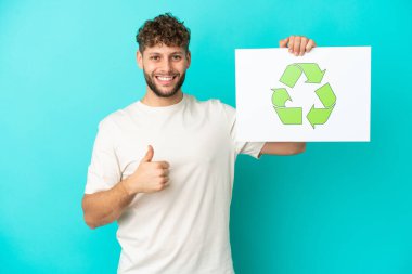 Mavi arka planda izole edilmiş yakışıklı beyaz bir adam elinde geri dönüşüm simgesiyle baş parmağıyla bir pankart tutuyor.