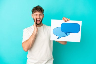 Genç beyaz adam mavi arka planda izole edilmiş. Elinde konuşma baloncuğu ikonu ve bağıran bir levha var.