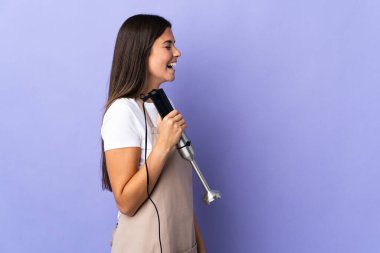 Mor arka planda izole edilmiş el blenderi kullanan Brezilyalı kadın yan pozisyonda gülüyor.