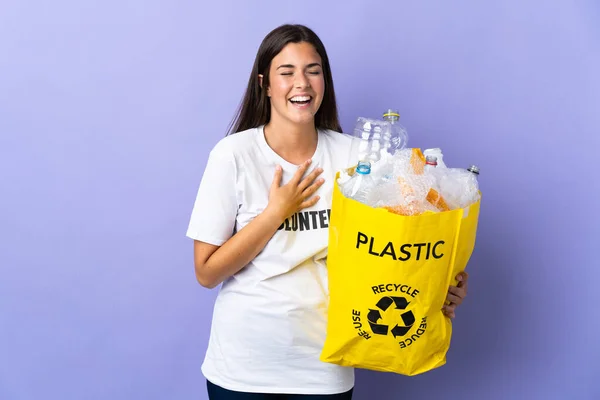 若いブラジル人女性が袋を持っていますプラスチックボトルでいっぱい紫色の背景に隔離されたリサイクル多くの笑顔 — ストック写真