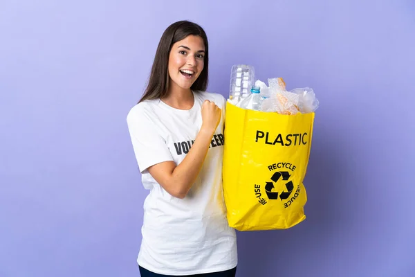 若いブラジル人女性は 勝利を祝う紫色の背景に隔離されたリサイクルするためにペットボトルでいっぱいの袋を持っています — ストック写真