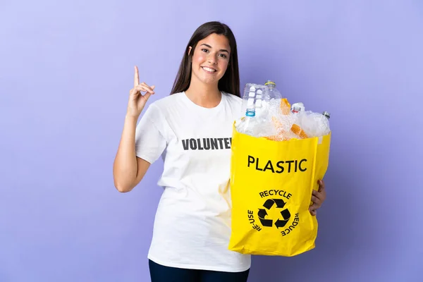 若いブラジル人女性は 紫色の背景を示す上で隔離されたリサイクルするためにビニールボトルでいっぱいの袋を保持し 最高の兆候で指を持ち上げる — ストック写真