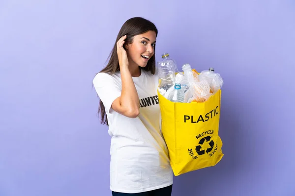 若いブラジル人女性は耳に手を置くことによって何かを聞く紫色の背景に隔離されたリサイクルするためにペットボトルの袋を持っている — ストック写真