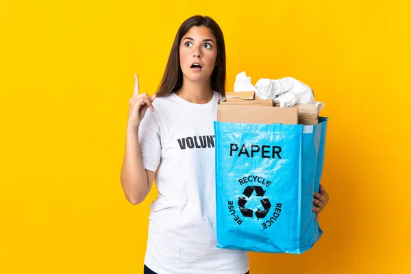 リサイクルバッグを持っている若いブラジル人の女の子紙でいっぱいリサイクルする黄色の背景に隔離された考え方指を指差す — ストック写真