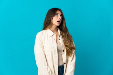 Genç beyaz kadın mavi arka planda izole edilmiş yukarı bakıyor ve şaşırmış bir ifadeyle