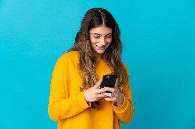Mavi arka planda izole edilmiş genç beyaz kadın cep telefonuyla mesaj ya da e-posta gönderiyor.