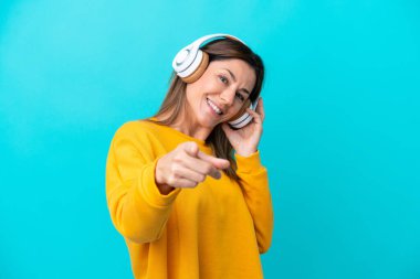 Orta yaşlı beyaz kadın mavi arka planda izole edilmiş müzik dinliyor ve ön tarafı işaret ediyor.
