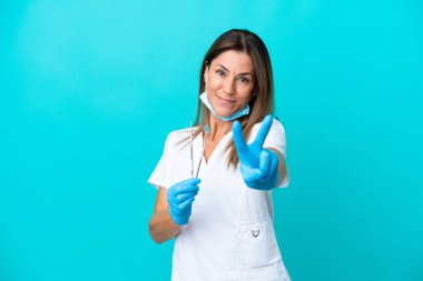 Mavi arka planda izole edilmiş orta yaşlı doktor kadın gülümsüyor ve zafer işareti gösteriyor.