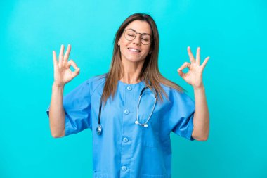 Orta yaşlı cerrah kadın Zen pozunda mavi arka planda izole edilmiş.