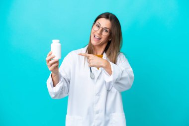 Mavi arka planda izole edilmiş orta yaşlı bir kadın doktor önlüğü giyiyor ve elinde haplar var.