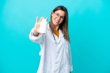 Mavi arka planda izole edilmiş orta yaşlı bir kadın doktor önlüğü giyiyor ve elinde haplar var.