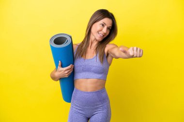 Elinde paspas tutarken yoga derslerine giden spor kadını sarı olmayan bir arkaplanda baş parmak işareti yapıyor.