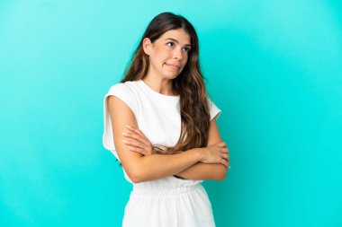 Mavi arka planda izole edilmiş genç beyaz kadın, omuzlarını kaldırırken şüpheyle hareket ediyor.