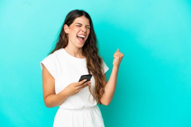 Genç beyaz kadın mavi arka planda izole edilmiş ve telefon zafer pozisyonunda.