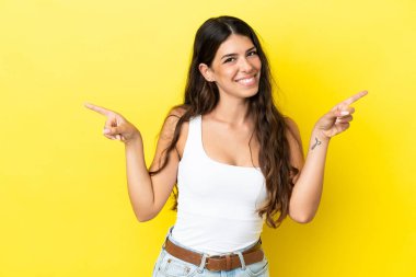 Sarı arka planda izole edilmiş genç beyaz kadın parmak uçlarını işaret ediyor ve mutlu.