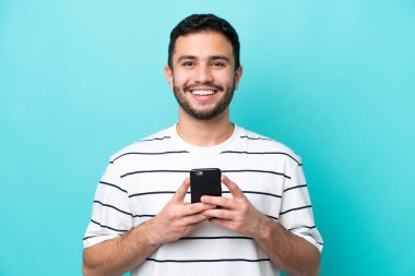 Genç Brezilyalı adam mavi arka planda izole edilmiş kameraya bakıyor ve cep telefonunu kullanarak gülümsüyor.