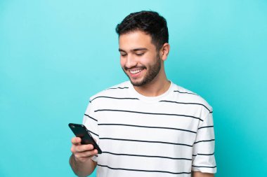 Genç Brezilyalı adam mavi arkaplanda izole edilmiş cep telefonuyla mesaj ya da e-posta gönderiyor