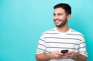 Genç Brezilyalı bir adam mavi arka planda cep telefonu kullanarak izole edilmiş.