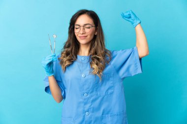 Mavi arka planda izole edilmiş aletleri tutan dişçi kadın güçlü bir hareket yapıyor.
