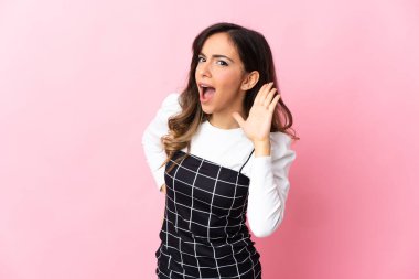 Pembe arka planda izole edilmiş genç beyaz kadın elini kulağına koyarak bir şeyler dinliyor.