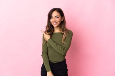 Pembe arka planda izole edilmiş genç beyaz kadın bir ürünü sunmak için kenarı işaret ediyor.