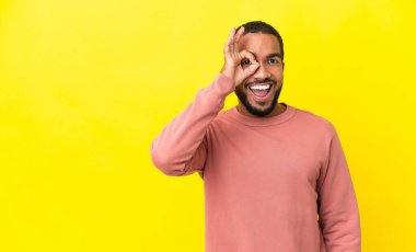 Sarı arka planda izole edilmiş genç Latin adam parmaklarıyla tamam işareti gösteriyor.