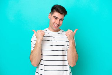 Kafkasyalı genç adam mavi arka planda izole edilmiş. Baş parmakları havada ve gülümsüyor.