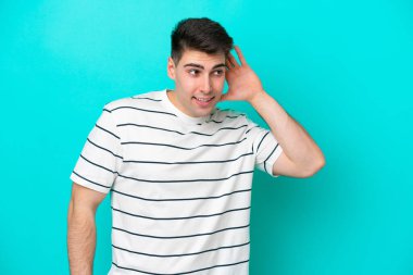 Kafkasyalı genç adam mavi arka planda izole edilmiş bir şekilde elini kulağına koyarak bir şeyler dinliyor.