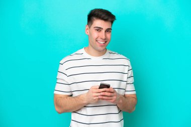 Mavi arka planda izole edilmiş genç beyaz adam cep telefonuyla bir mesaj gönderiyor.