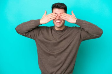Mavi arka planda izole edilmiş genç beyaz bir adam elleriyle gözlerini kapatıyor ve gülümsüyor.