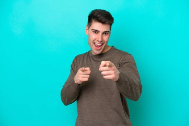 Mavi arka planda izole edilmiş genç beyaz adam ön tarafı işaret ediyor ve gülümsüyor.