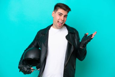 Motosiklet kasklı genç beyaz adam mavi arka planda izole edilmiş, şok olmuş yüz ifadesi var.