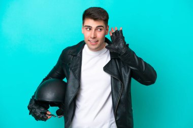 Motosiklet kasklı genç beyaz adam mavi arka planda izole edilmiş bir şekilde elini kulağına koyarak bir şeyler dinliyor.