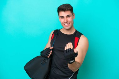 Spor çantalı beyaz tenli genç bir adam mavi arka planda izole edilmiş bir şekilde bizimle gelmesi için davet ediliyor. Gelmene sevindim.